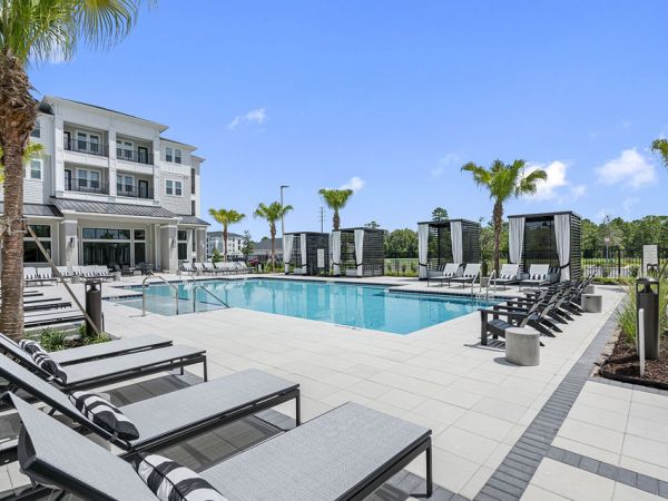 Modern outdoor pool area with lounge chairs, palm trees, and cabanas beside a multi-story apartment building.