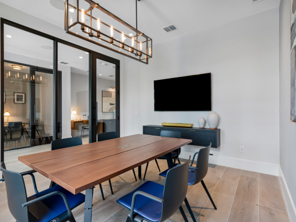 Modern conference room with a wooden table, six chairs, wall-mounted TV, and large glass doors.