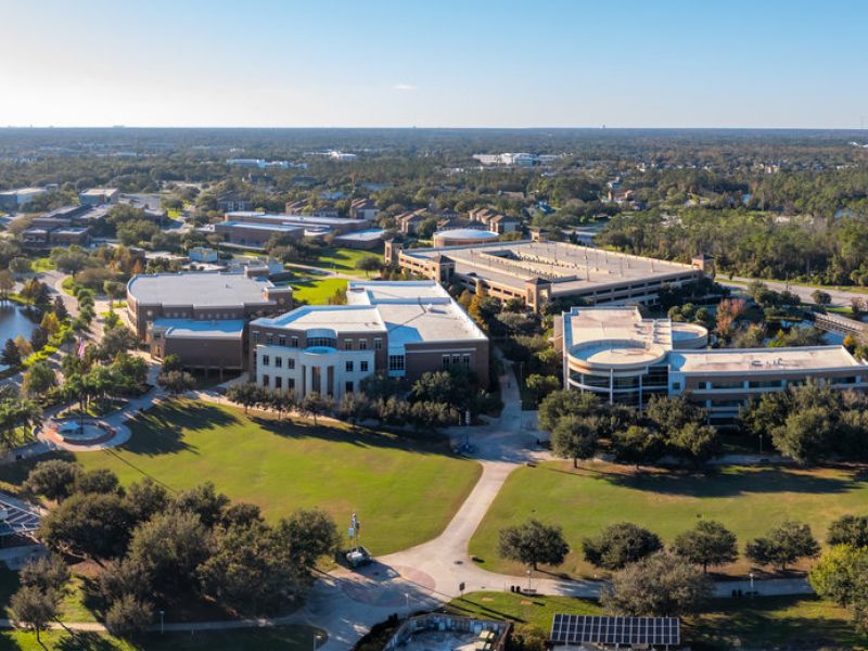 Aerial view of a university campus with modern buildings, green lawns, and surrounding trees under a clear sky.