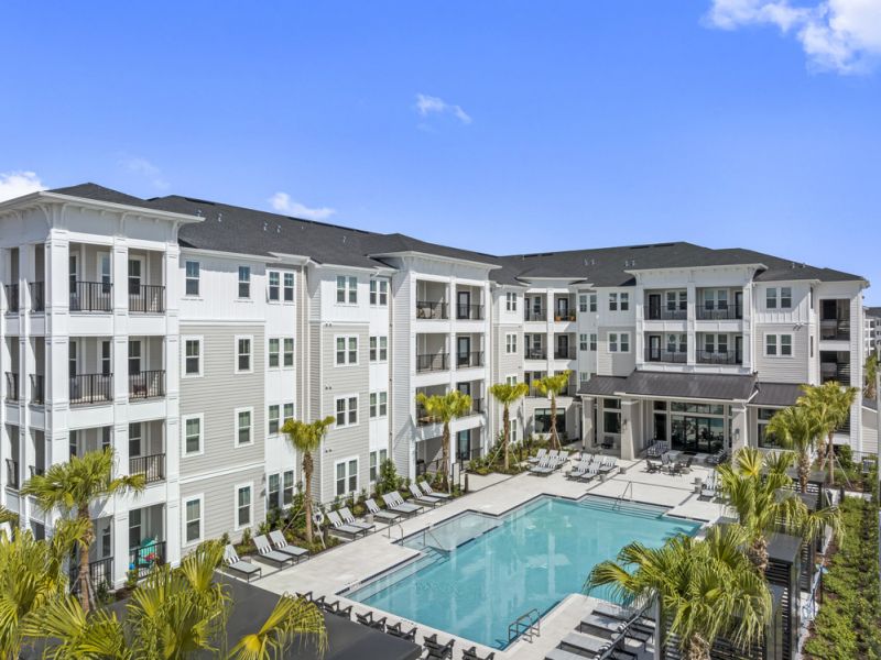 A modern four-story apartment complex with balconies overlooks a large outdoor swimming pool and palm trees.