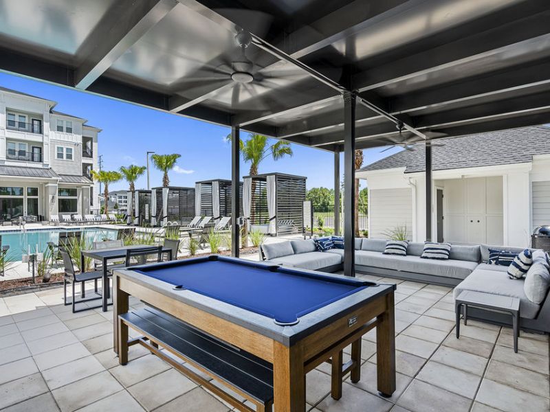 Outdoor lounge with pool table, modern sofa seating, and a pool area in the background, under a covered patio.