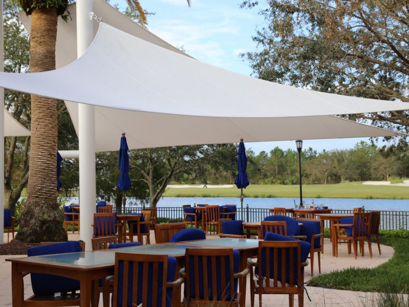 Outdoor patio with wooden tables, blue chairs, and sun shade sails, overlooking a lake and golf course.