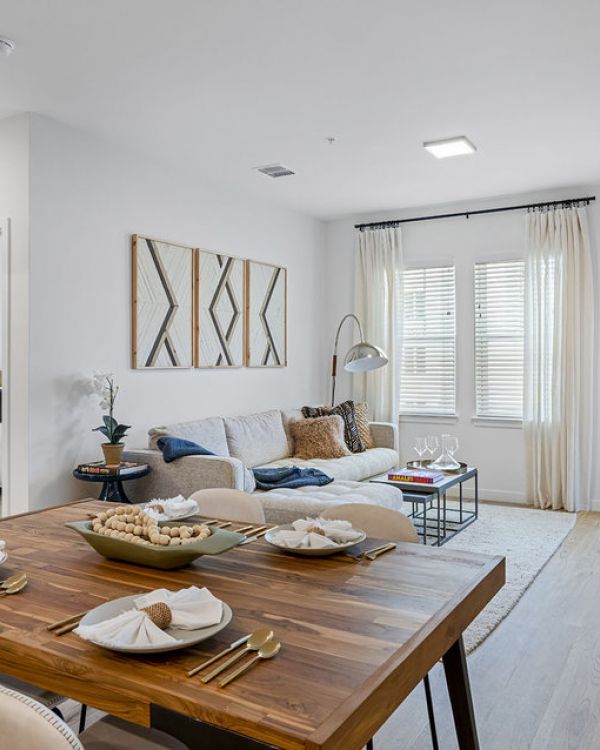 Modern living and dining area with a wooden table, white chairs, sofa, and large windows with curtains.