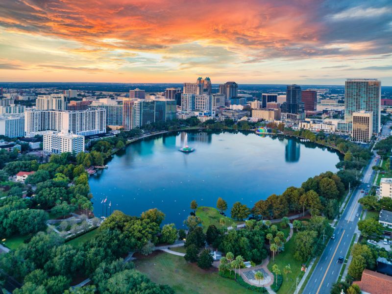 Aerial view of a city skyline at sunset with a large circular lake surrounded by trees in the foreground.