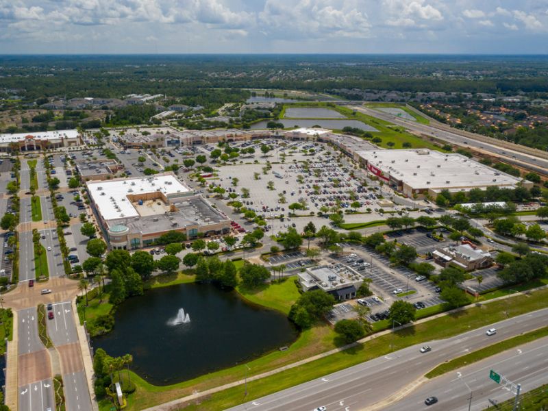 Aerial view of a large shopping center with parking lots, nearby roads, and a pond with a fountain.